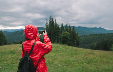 Back view of a female traveler in a red jacket using her smartphone and taking photos of a beautiful mountain scenery: mountain hills and woods.の写真素材