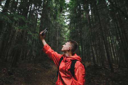 Young backpacker guy in a bright red raincoat taking selfie on his smartphone in the mountain woods....