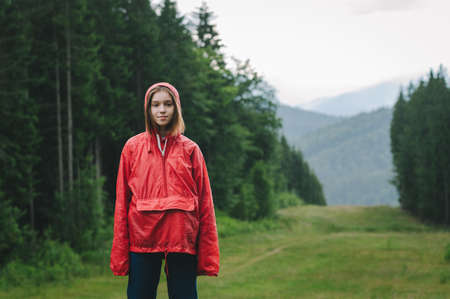 Young pretty tourist female is posing for the photo on a mountain hill in a red waterproof jacket under the summer rain. Wet weather.の写真素材