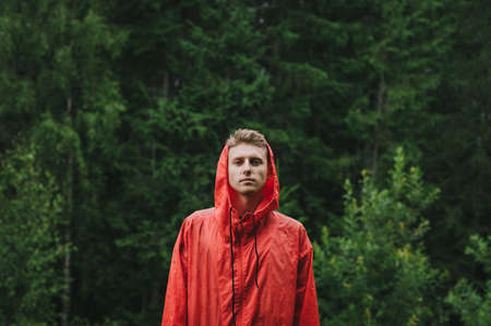 Portrait of a young pensive calm male tourist standing in front of the green mountain forest in the pouring rain wearing a red waterproof jacket, feeling peaceful and calm.の写真素材