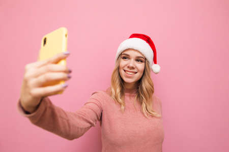 Positive girl in pink sweater and santa hat stands on pink background and poses on smartphone camera...