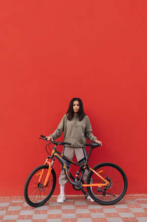 Cheerful woman in sportswear stands with a bicycle on a background of a red wall and poses for the camera. Cycling down the street.の写真素材