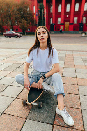Stylish woman in casual clothes sits on a longboard outdoors after skiing, relaxing and looking at...