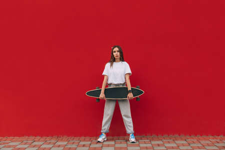 Stylish woman in street casual clothes posing at camera with a longboard in her hands on a red wall background. Portrait of a skater woman on a red background.の写真素材