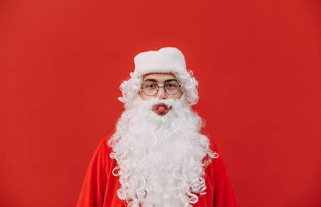 Funny Santa stands on a red background, looks at the camera and shows his tongue. Funny santa claus with beard makes funny face isolated on redの写真素材