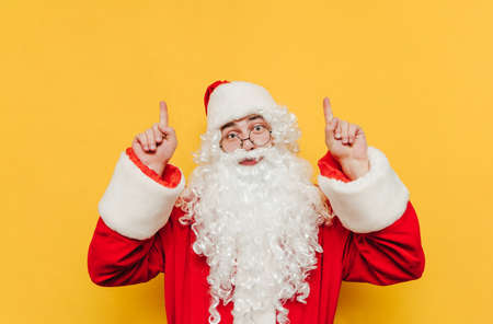 Funny Santa Claus showing thumbs up on copy space with serious face looking at camera, isolated on yellow background. Christmas concept.の写真素材