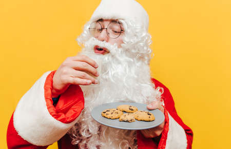 Funny santa claus drinks milk and eats chocolate christmas cookies on yellow background, close up photo. New Year.の写真素材