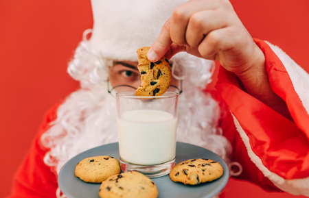 Santa Claus dips Christmas cookies in a glass of milk, close-up cropped photo. Background. Santa eats cookies at Christmasの写真素材