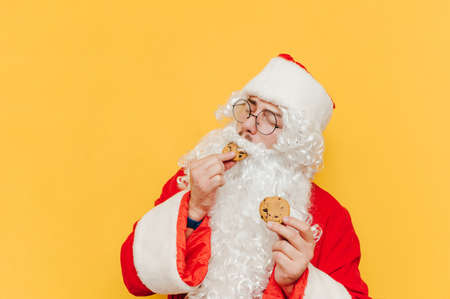 Portrait of happy santa claus eating sweet chocolate chip cookies with closed eyes on yellow background. Santa eating christmas sweets, isolated.の写真素材