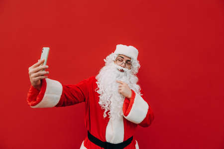 Santa Claus taking selfie on smartphone on red wall background. Christmas and New Year selfie.