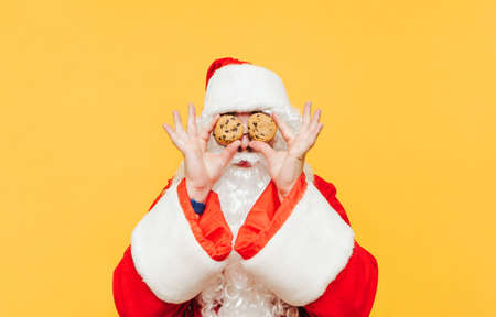 Funny santa with chocolate chip cookies in hands holding eyes near yellow background. Santa Claus and Christmas cookies, isolated. New Yearの写真素材