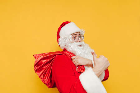 Positive Santa with a bag stands on a yellow background, shows a thumb up and looks at the camera with a smile on his face. Christmas concept.の写真素材