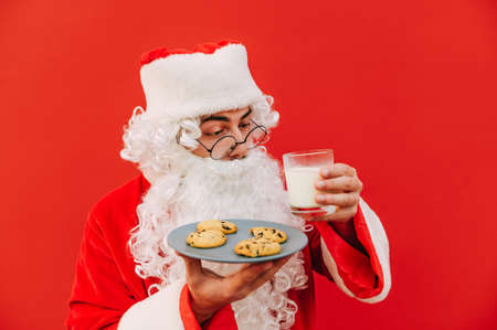 Funny santa isolated on red background with a glass of milk and a plate of chocolate chip cookies. Christmas and New Year concept.の写真素材