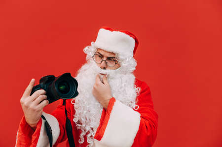 Santa Claus isolated on red background with a camera in his hands, looking with a pensive face. Christmas photographer.の写真素材