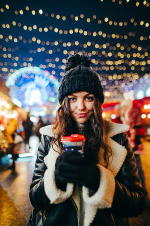 Close-up evening portrait of a cute girl at the Christmas market, standing with a cup of warming drink on the background of a decorated fair with flashlights. Lady at the Christmas market. Verticalの写真素材