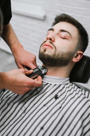 Barber shaves an electric beaver shaver in a barber shop chair. Male percussionist does hairstyle to client, holds trimmer in hands. A man lies in a chair and cuts a beard in a barber.の写真素材