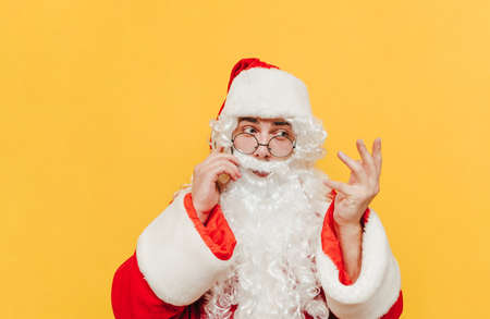 Attractive Santa Claus is talking with someone via mobile phone, having a discussion, gesticulating, raising his hand, trying to explain. Santa is standing at the studio on a yellow background.の写真素材