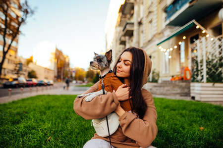 Beautiful woman in casual clothes sits on the grass on the street background with a biewer terrier in her arms, looks to the side and smiles. Owner sits on the lawn with a pet on a street background.の写真素材