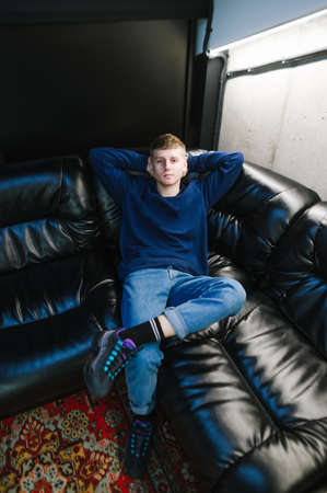 Attractive young man in casual clothes and stylish sneakers resting on a black leather sofa in a dark room, looking into the camera with a serious face. Vertical.の写真素材