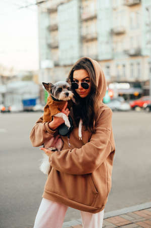 Portrait of a stylish woman in casual clothes on a background of cityscape standing on the street with a small dog in her arms, looking at the camera with a serious face and posing. Verticalの写真素材