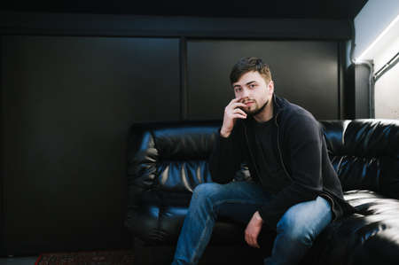 Adult man with a beard and casual clothes sits on a leather sofa in a dark room with a black wall, posing for a camera with a serious face. Man relaxing sitting on black couch, looking into camera.の写真素材
