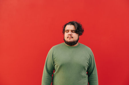 Young bearded fat hispanic man with a stylish haircut is looking down with suspicion, standing in front of the red wall, feeling confused.の写真素材