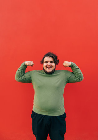 Vertical shot of a strong young fat hispanic man demonstrating his power by raising his hands and clenching his fists, showing muscles, smiling and standing on a red background.の写真素材
