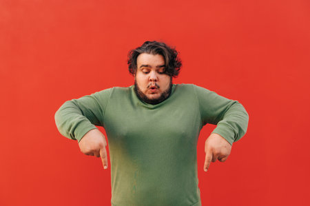 Funny obese hispanic guy with a stylish haircut is pointing his fingers down, advertising a product, being emotional, standing in front of the red wall.の写真素材