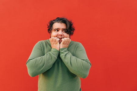 Scared young overweight hispanic guy with a stylish haircut is standing in front of the red wall,...