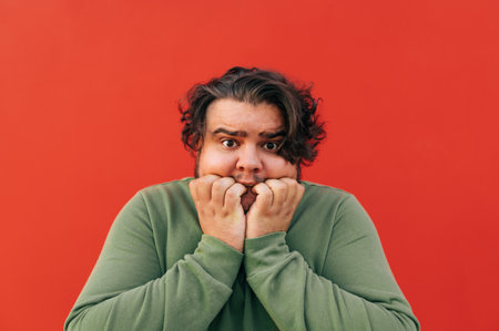 Closeup portrait of a scared young corpulent guy with a curly hair standing on a red background, touching his face with a frightened expression, feeling terrified.の写真素材