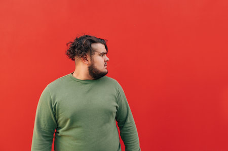 Side view of an overweight bearded hispanic young man looking aside, being serious and confident, standing on an isolated red background.の写真素材