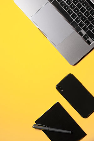 Vertical flat lay composition with a silver gray laptop, black smartphone, notepad and a pen on an...