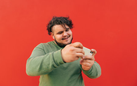Focused overweight hispanic guy is enjoying playing games on his smartphone, being concentrated on the process, smiling and willing to win.の写真素材