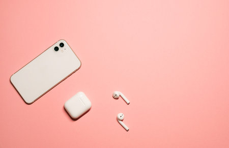 Simple flat lay photo on a pastel pink background with a white smartphone and wireless earphones...