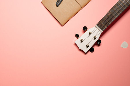 Creative layout with a wooden white ukulele guitar fretboard with nylon strings, brown notebook, pen, and a white pick on a pastel pink background.の写真素材