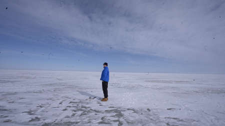 A man in a blue jacket stands on a frozen lake and looks into the distance.の写真素材