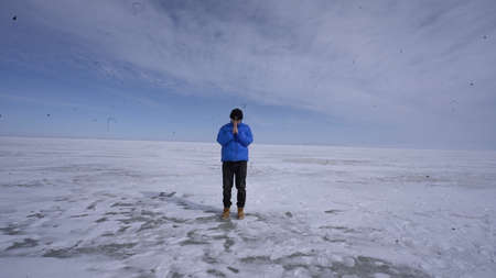 A man in a blue jacket stands in the middle of a frozen lake and looks at the horizon.の写真素材