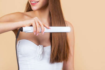 Smiling woman ironing long hair with flat iron. Happy young woman straightening hair with straightener. Portrait of young beautiful girl using styler on her shining hair. Hairstyleの写真素材
