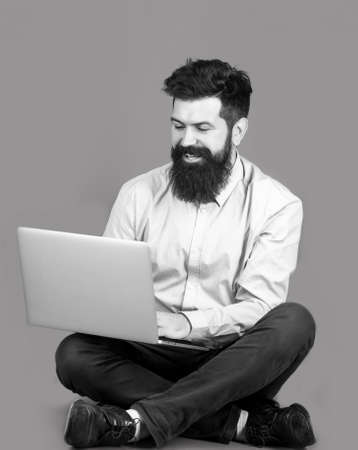 Smiling handsome bearded man worker laptop. Happy young man sitting on the floor with and using laptop computer on red background. Young businessman using his laptop, pc. Black and whiteの写真素材