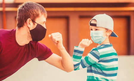 Social distancing concept. Coronavirus outbreak. Protective measures. Father puts her son a face protective mask outdoors. Father and son bump elbows outdoors. Coronavirus quarantineの写真素材