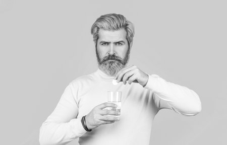 Male taking a pill with a glass of water. Man take some pills, holds glass of water, isolated on blue. Black and white. Berded man taking drugs to releave headache. Man taking pill against headacheの写真素材