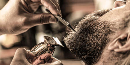 Barber works with a beard clipper. Hipster client getting haircut. Hands of a hairdresser with a beard clipper, closeup. Bearded man in barbershop. Man visiting hairstylist in barbershopの写真素材