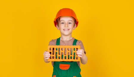 Boy in a construction helmet holds a brick in his hands on yellow background. Little builder in helmet. Child dressed as a workman builder. Little boy wearing helmet. Child building helmetの写真素材