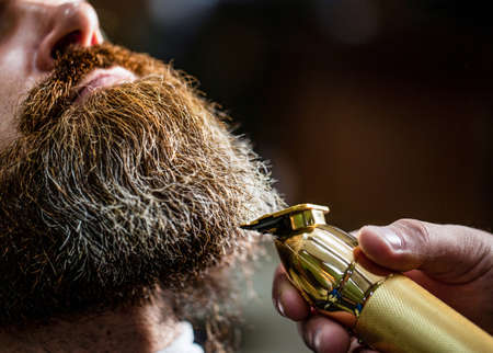 Man visiting hairstylist in barbershop. Barber works with a beard clipper. Hipster client getting haircut. Hands of a hairdresser with a beard clipper, closeupの写真素材