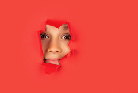 Spy watching through hole. Boy peeks through a cut hole in red paper wall. Positive child with smile on face. Through paper. Little boy looking through paper.の写真素材