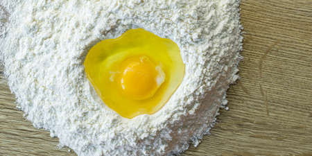 A yellow egg is smashed in a heap of flour on a wooden table. Make a dough.