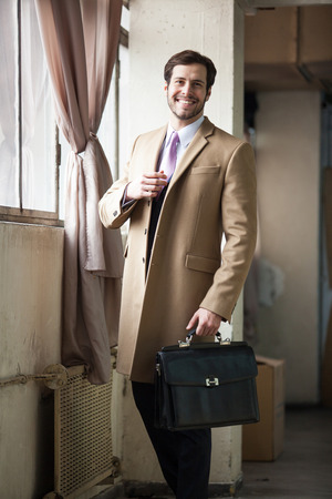 Friendly and smiling businessman looking at camera, wearing beige coat, white chemise, pink tie, black trousers and holding his black briefcase.の写真素材