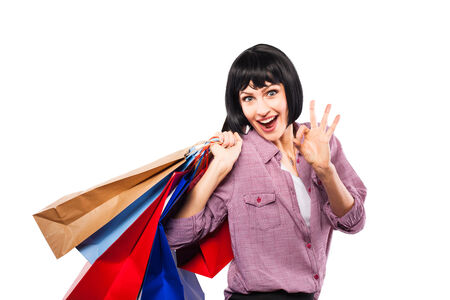 happy young brunette woman smiling and showing ok sign with shopping bags over her shoulder, on white backgroundの写真素材