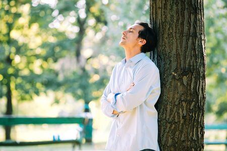 adult man in blue shirt leaning on a tree in parkの写真素材
