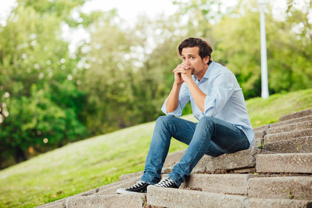 adult man in blue shirt sitting alone on stairs outside and thinkingの写真素材